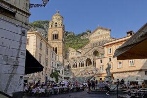 Piazza Duomo ad Amalfi affollata di visitatori: le terrazze dei bar occupano gran parte dello spazio aperto davanti alla scalinata della cattedrale.